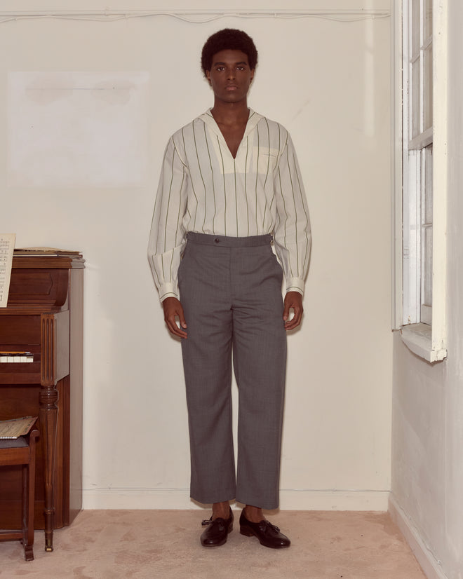 A person stands indoors in a striped shirt, midcentury style with Tropical Wool Side-Buckle Trousers in grey, and black shoes next to a piano.