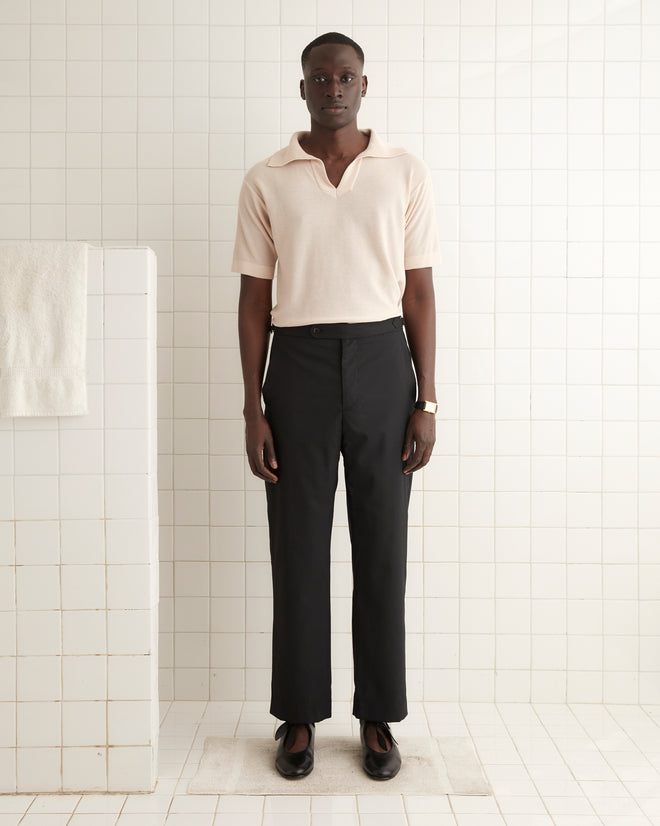 A person stands in a tiled room, wearing a light polo shirt and Tropical Wool Side-Buckle Trousers in black, with a watch on their wrist.