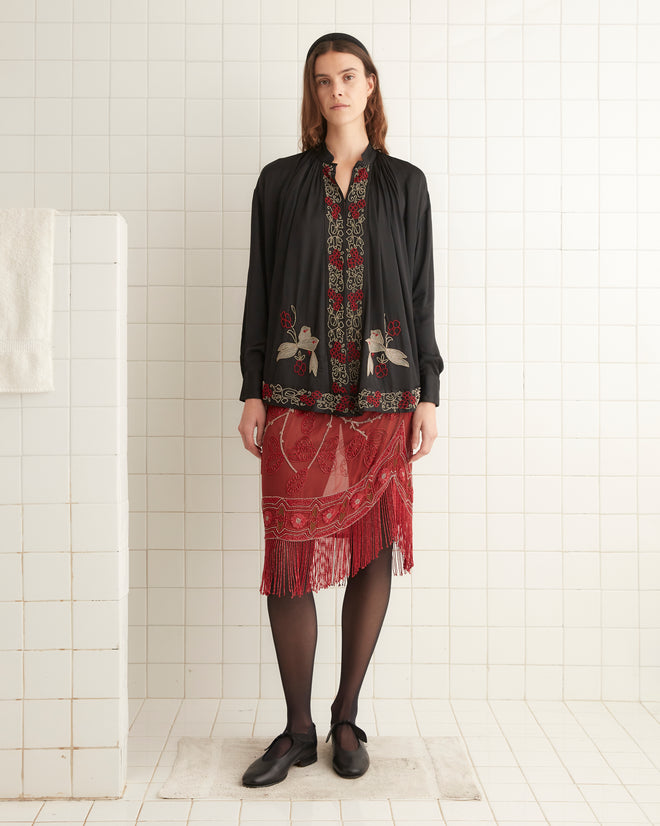 A woman stands in a tiled room wearing a Beaded Tropicbird Shirt adorned with pearl buttons, a red patterned skirt with fringe, black tights, and black shoes.