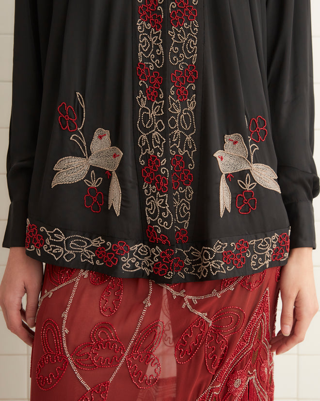 A woman stands in a tiled room wearing a Beaded Tropicbird Shirt adorned with pearl buttons, a red patterned skirt with fringe, black tights, and black shoes.