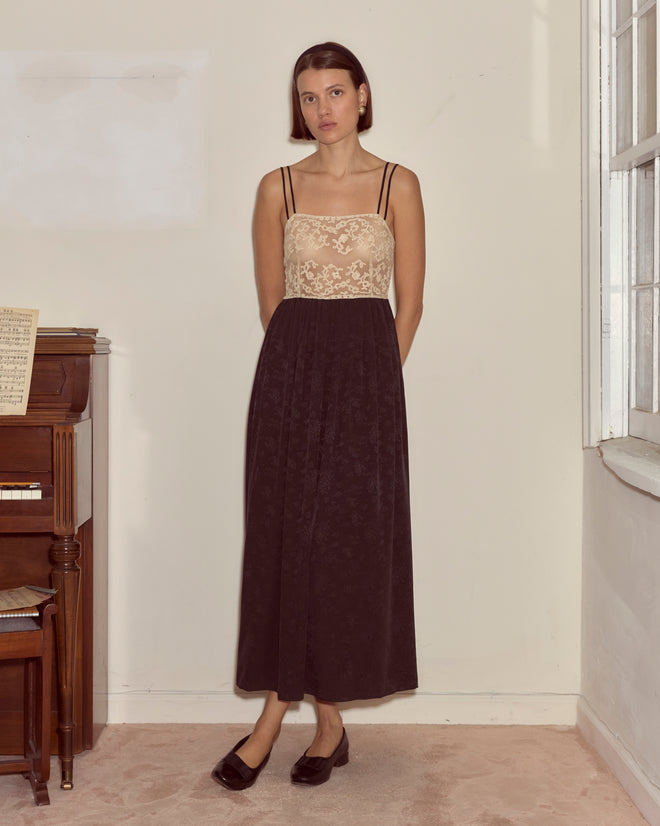 A woman in the Trousseau Lace Slip Dress - Black stands indoors by a piano and window, hands behind her back, gazing ahead.