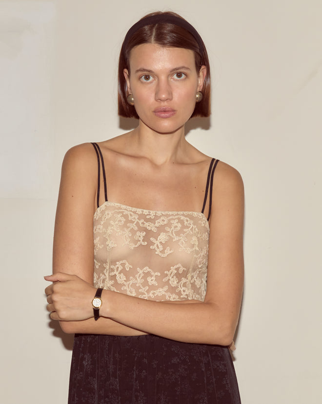 Woman with short brown hair wears the Trousseau Lace Slip Dress in black, paired with pearl earrings and a watch, standing against a plain wall.