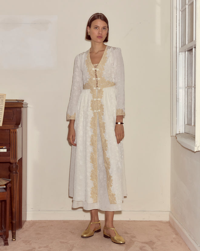 A woman in a Trousseau Lace Dress - Cream stands indoors near a piano and a window, looking at the camera.