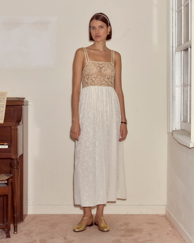 A woman in the Trousseau Lace Slip Dress - Cream, featuring sheer silk and a sleeveless design, stands indoors by a piano near a sunlit window.