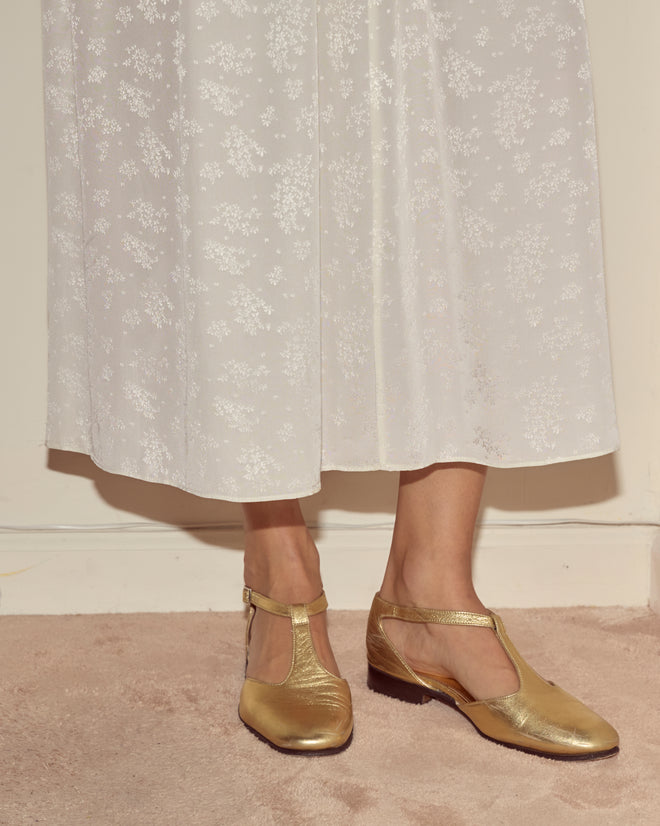 A person wearing the Trousseau Lace Slip Dress in cream and gold T-strap shoes stands on a beige carpet.