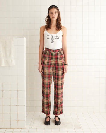 A person with long hair stands in a tiled room wearing a white tank top with 