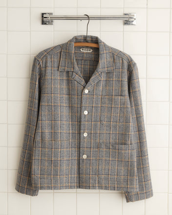 The Tumbleweed Plaid Overshirt - M, with its gray plaid pattern that echoes 1960s suiting fabric and is made from 100% wool, hangs on a hanger against a white tiled wall.