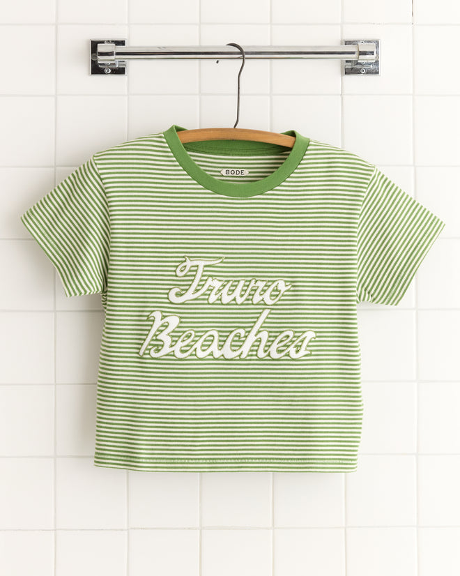 A 1950s-inspired Truro Stripe Cropped Tee, featuring green and white stripes with "Truro Beaches" text, made from 100% cotton, hanging on a rod against a white tiled wall.