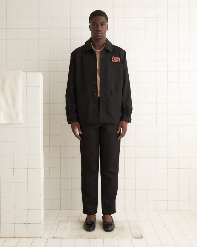 A man stands in a white tiled room, facing the camera. He is wearing a black Twill Franck Jacket, black pants, a beige shirt underneath, and black shoes. A white towel hangs on a rack to his left.