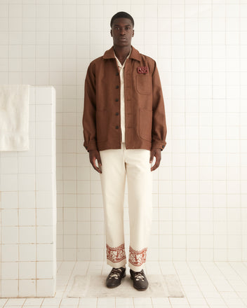 A man stands against a white tiled wall wearing the Twill Franck Jacket in brown, cream pants with intricate designs at the hem, and dark shoes. A white towel hangs on a tiled structure beside him, evoking the timeless charm of 1940s men's coat styles.