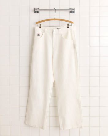 The Twill Knolly Brook Trousers in white, featuring a vintage workwear silhouette, hang from a wooden hanger against a white tiled wall.