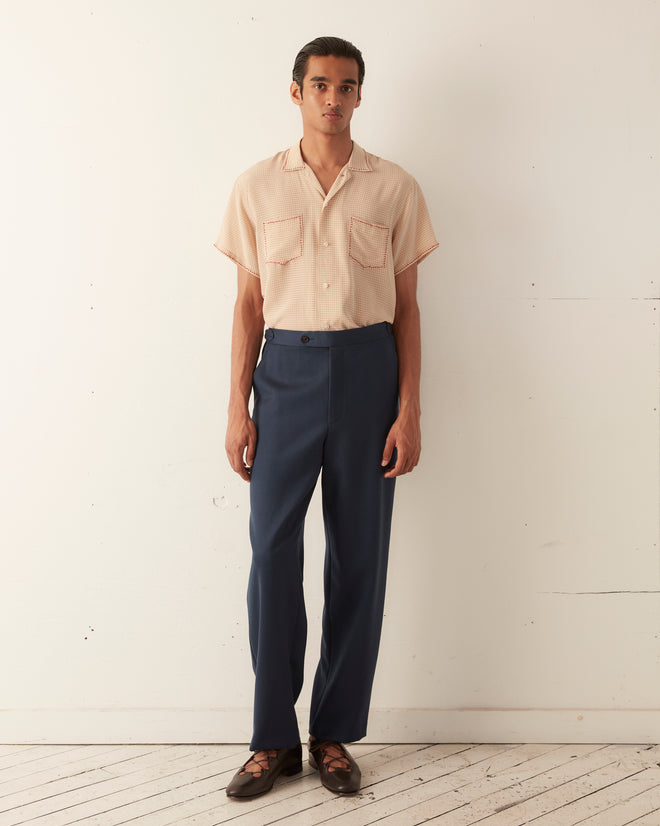 Someone in a peach shirt and Twill Side Buckle Trousers - Petrol stands against a plain white wall.