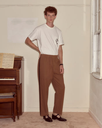 A man stands indoors by an upright piano, wearing a white t-shirt, black shoes, and Twill Track Pants - Brown.