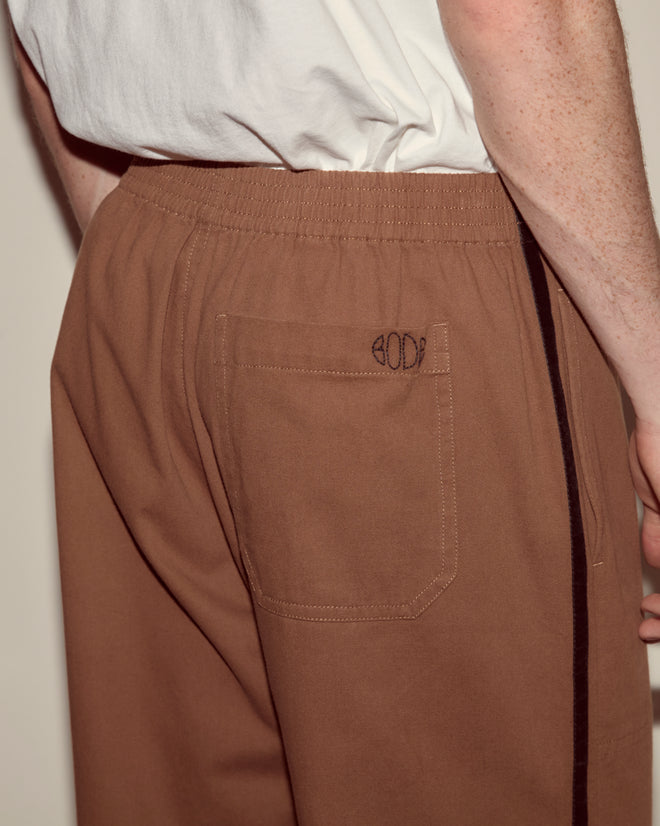 A person wears Twill Track Pants - Brown featuring heavyweight cotton twill, a back pocket, and "BODE" stitched above the pocket.