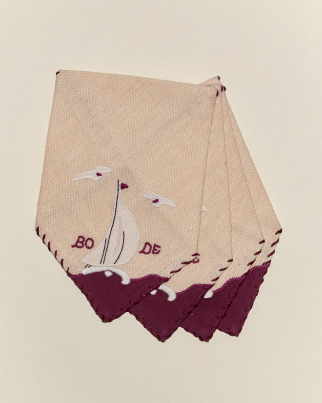 Set of four tan linen Twin Sails Cocktail Napkins with maroon edges, each featuring a white sailboat appliqué and embroidered with “BORD DE”.