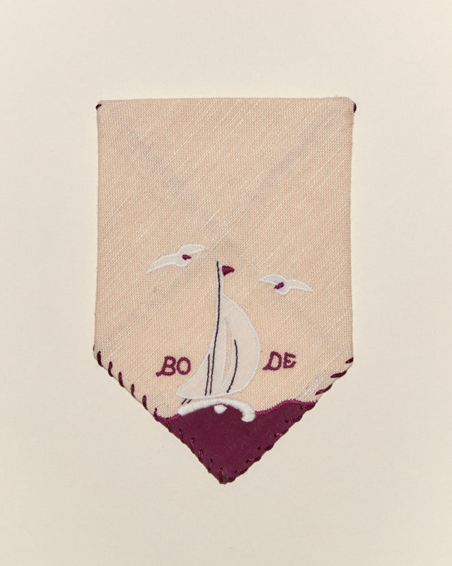 The Twin Sails Cocktail Napkins feature a cream fabric patch with a sailboat appliqué, two birds, and "BODE" embroidered in purple—ideal for accenting linen or cocktail napkins.