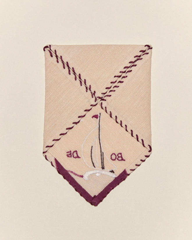 The Twin Sails Cocktail Napkins feature beige fabric with dark purple stitching, embroidered with a white bird and "BO DE," offering a charming, classic linen napkin accent to your table.
