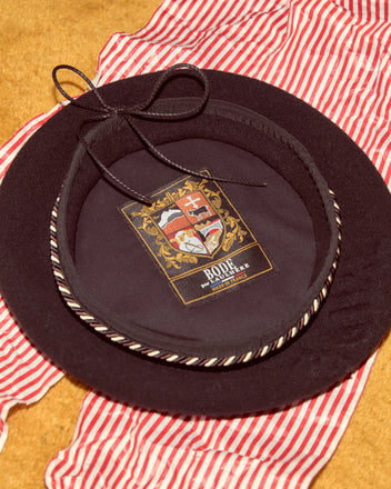 The Script Beret in black with brown/black piping, featuring a crest label inside and made in France, is shown atop red-and-white striped fabric.