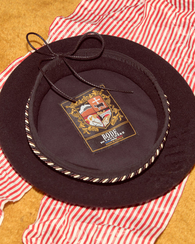 The Script Beret in black with brown/black piping, featuring a crest label inside and made in France, is shown atop red-and-white striped fabric.