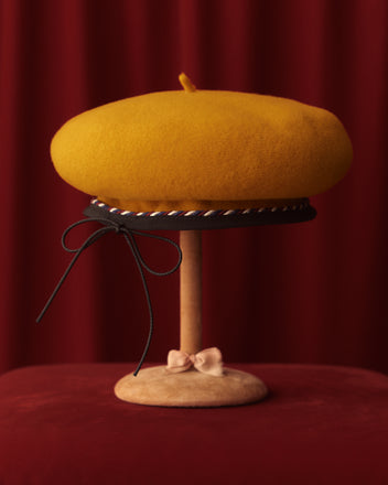 The Twisted Piping Beret - Golden, crafted from 100% wool, sits elegantly on a stand, its yellow hue contrasting beautifully against a deep red curtain backdrop.