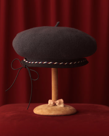 The Twisted Piping Beret in charcoal, crafted in France from 100% wool, is elegantly displayed on a wooden stand against a rich red velvet background and adorned with a decorative ribbon.