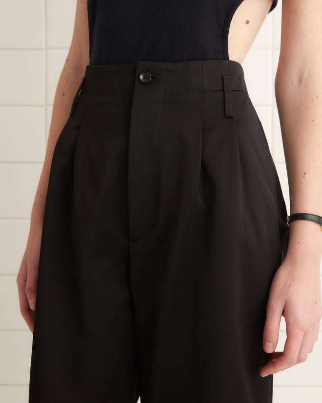 Wearing a black tank top and Tuxedo Braid Murray Trousers, crafted in a chic high-waisted, wide-leg style, the individual stands confidently against a tiled wall.