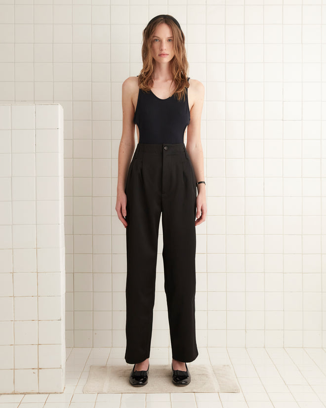 Wearing a black tank top and Tuxedo Braid Murray Trousers, crafted in a chic high-waisted, wide-leg style, the individual stands confidently against a tiled wall.