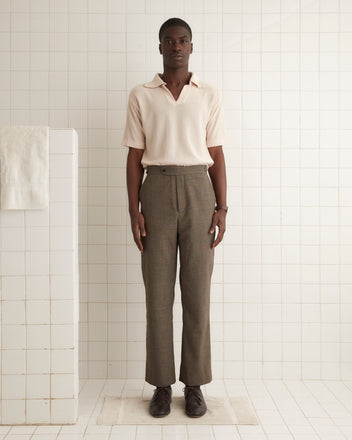 A person dressed in the beige Ume Glen Trousers - 31, crafted from vintage suiting fabric, stands in a tiled room, facing forward with a neutral expression.