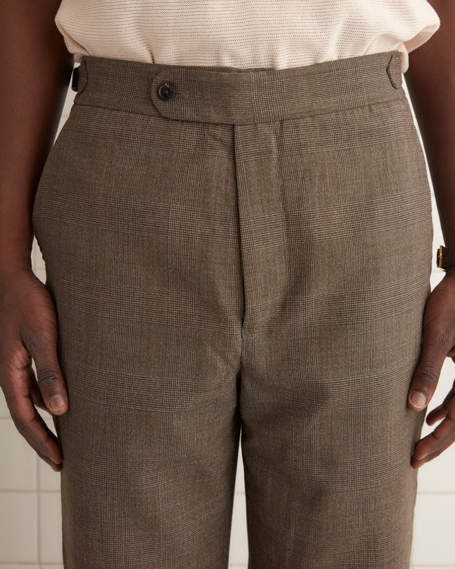 A person dressed in the beige Ume Glen Trousers - 31, crafted from vintage suiting fabric, stands in a tiled room, facing forward with a neutral expression.