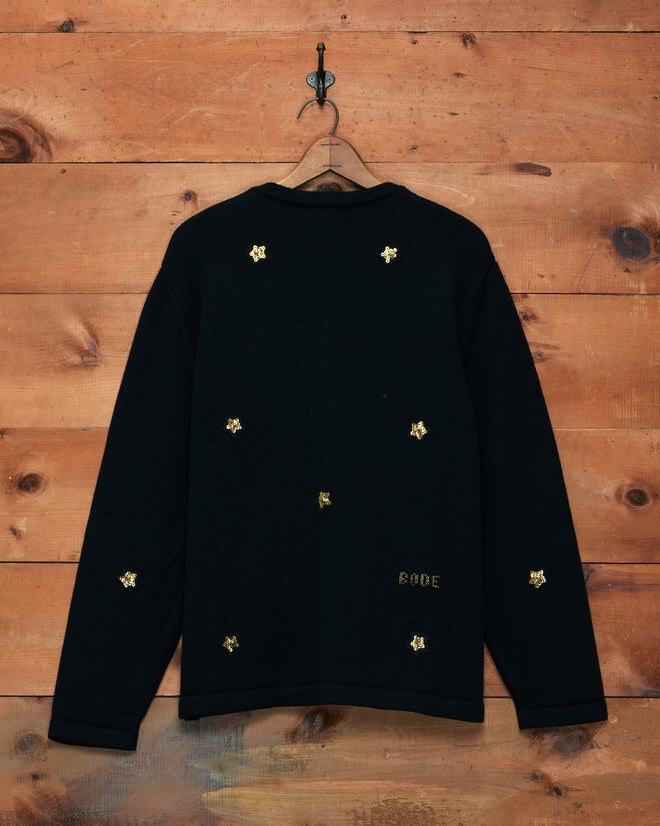 Star Pony Cardigan—a black sweater adorned with small gold embroidered designs scattered across the back, perfect for those who appreciate refined men’s knitwear—displayed hanging on a wooden wall.