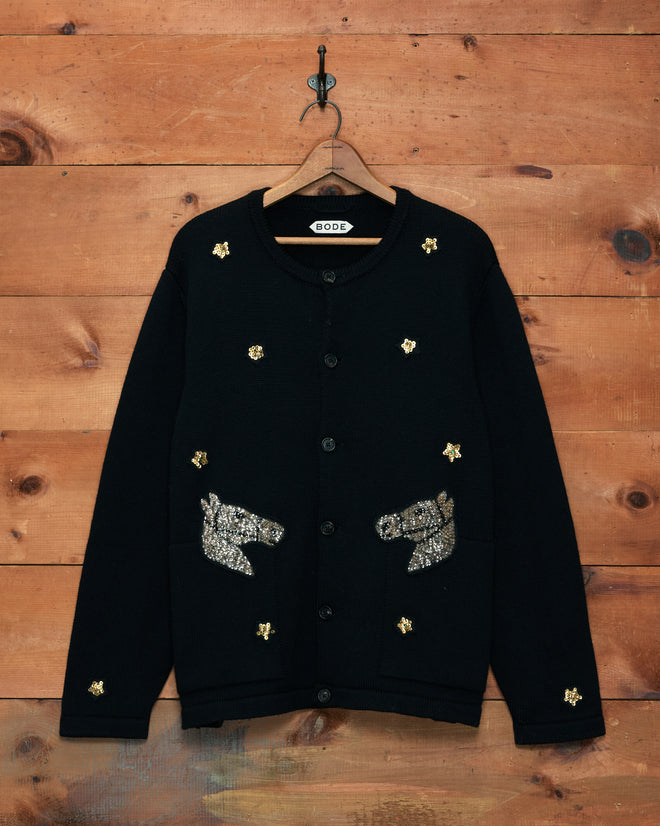 The Star Pony Cardigan—a black virgin wool men’s knitwear piece—features horse head and star sequin patches, displayed on a wooden wall with a hook for standout style.