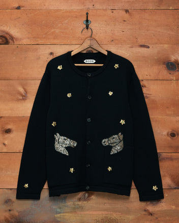 The Star Pony Cardigan—a black virgin wool men’s knitwear piece—features horse head and star sequin patches, displayed on a wooden wall with a hook for standout style.