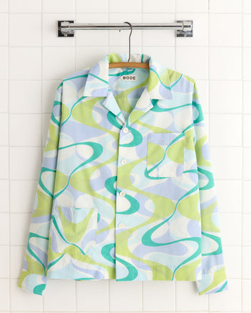 The Upstream Creek Shirt, a unique button-up with a blue, green, and white abstract pattern, hangs on a hanger against a tiled wall.