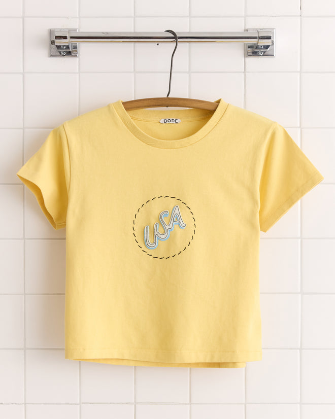 The USA Tee is a yellow short-sleeve shirt made of 100% cotton, featuring "USA" text inside a dashed circle, displayed on a hanger against a tile wall.