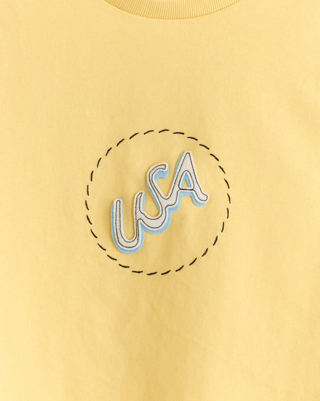 The USA Tee is a retro-inspired yellow 100% cotton shirt with "USA" in blue and white letters inside a dotted stitched circle, giving vintage 1970s vibes.