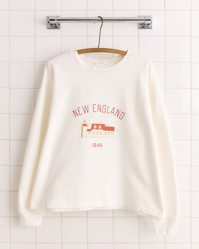 The New England Crewneck - White is a boxy-fit souvenir sweatshirt with "NEW ENGLAND," "1945," and a building graphic screen-printed on it, displayed on a wooden hanger.