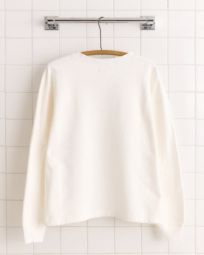 The New England Crewneck - White, a boxy-fit long-sleeve shirt, hangs on a wooden hanger against a white tiled wall.