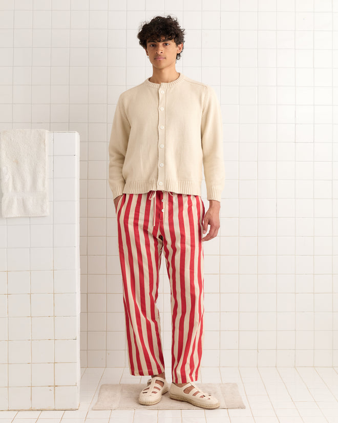 A man stands in a white tiled room wearing Valance Stripe Pajama Pants, a cream sweater with pearl buttons, and cream sandals.