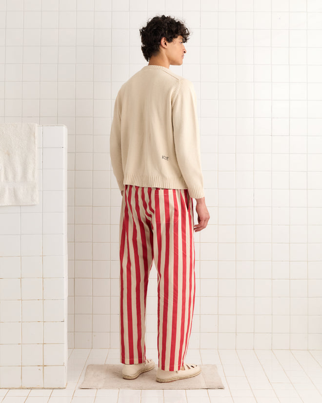 Wearing Valance Stripe Pajama Pants, a person in a cream sweater with pearl buttons stands on a mat in a tiled room, facing away—a look inspired by 1940s curtain valance.