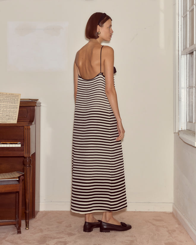A woman wearing the Velvet Bannister Dress stands sideways by a piano, gazing out a window in a softly lit room.