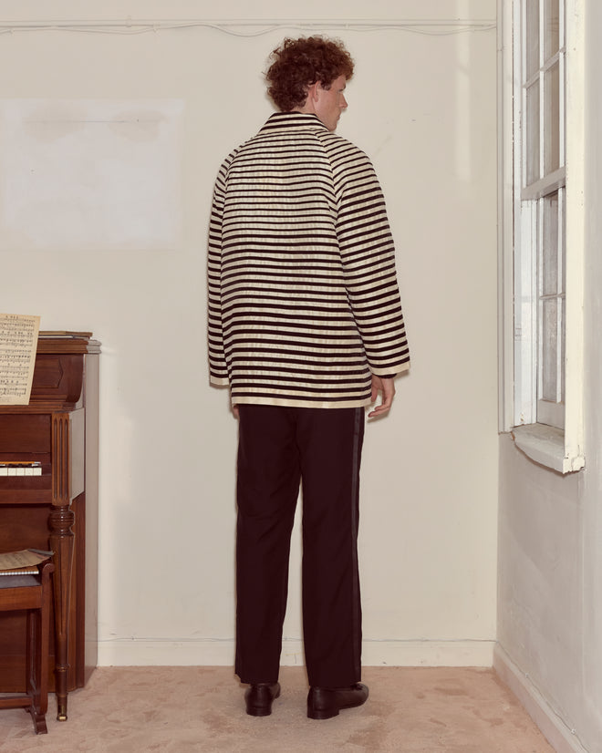 A person in a Velvet Bannister Jacket and black pants stands facing a blank wall in a room featuring a piano.