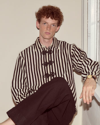 A person with curly hair wears the black and white Velvet Bannister Shirt, sitting by a window and looking at the camera.