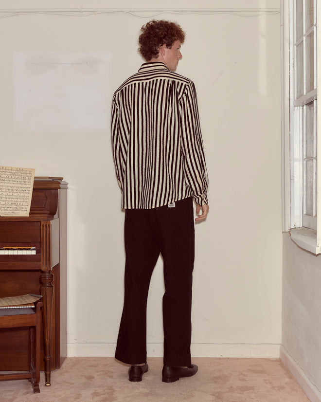 Wearing the Velvet Bannister Shirt, a black and white striped 100% silk top, and black pants, a person stands facing away in a room with a piano.
