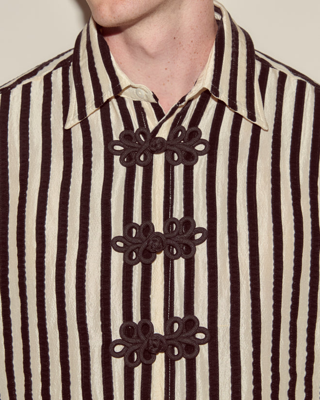A person wearing the Velvet Bannister Shirt, a black-and-white striped 100% silk shirt with velvet ribbon details and decorative black frog closures.