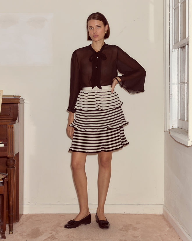 Woman in a sheer black blouse and Velvet Bannister Skirt stands indoors near a piano and window.