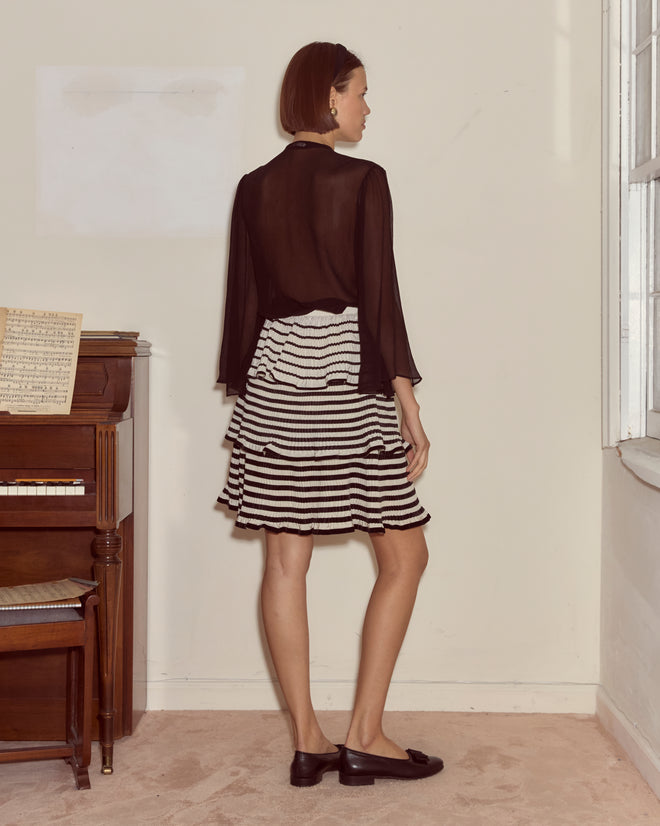 A woman stands by a piano in a softly lit room, facing away, wearing a black sheer top and the Velvet Bannister Skirt.