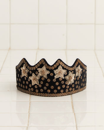 The Velvet Sequin Crown - Black, adorned with gold bullion stars and dot patterns, rests elegantly on a white tile background.