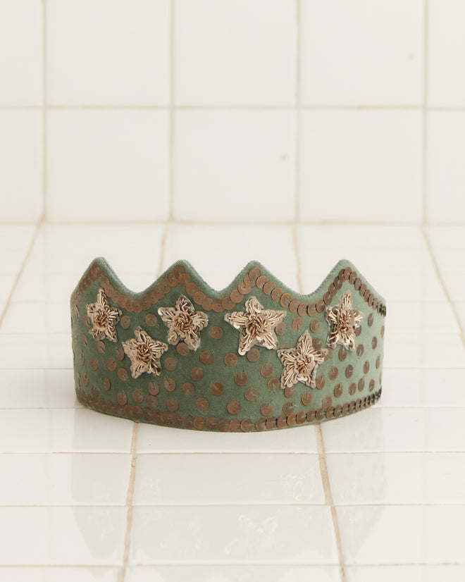 The Velvet Sequin Crown in a sage hue, embellished with gold bullion stars, is elegantly displayed on a tiled surface.