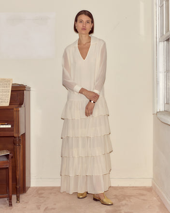 A woman in the Vendome Gown—a long, tiered silk dress with French cuffs—stands by a piano in a softly lit, neutral-toned room.