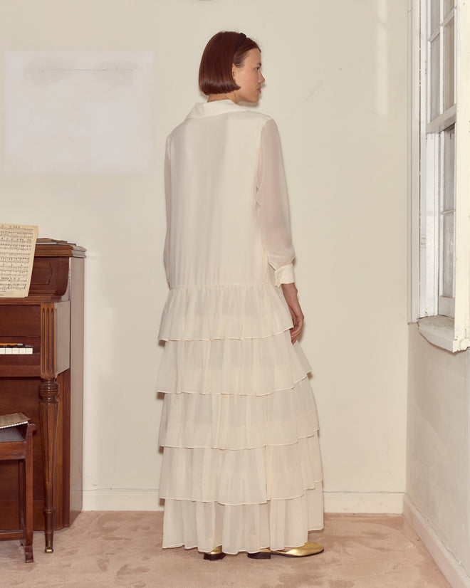 A woman wearing the Vendome Gown, a long white dress with a drop waist, stands by a piano facing away near a sunlit window.